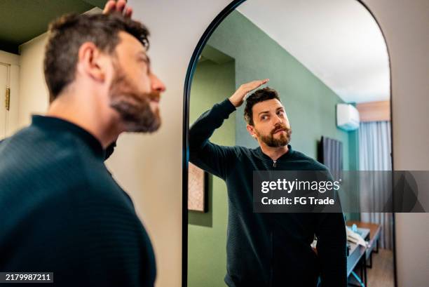 mature man fixing his hair in front of the mirror in a bedroom - hand in hair stock pictures, royalty-free photos & images