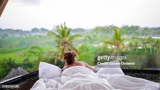 a woman enjoys a relaxing morning in a hammock with a view of rice fields and palm trees on the indonesian island of bali, a young woman lies in a netted hammock on the terrace of a bamboo hotel in bali and takes in the view - covers tour stock pictures, royalty-free photos & images