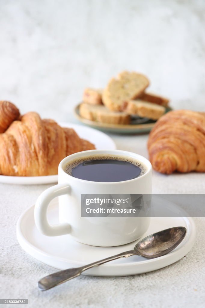 Nahaufnahme einer weißen Kaffeetasse aus Porzellan und einer Untertasse voller schwarzem Kaffee, Americano-Kaffee (Espresso und Wasser), Teelöffel, Tellern mit Biscotti (italienische Mandelkekse) und frisch gebackenem Frühstücksgebäck, goldenen Butter