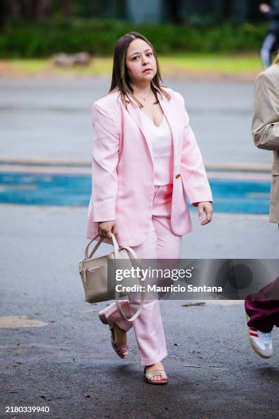 Guest wears pink blazer, matching pink trousers, white blouse, beige handbag, open-toe pink sandals, gold button detail, straight hair, minimal...