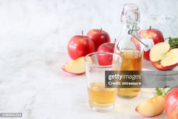 image of open, swing stopper bottle containing apple cider vinegar by drinking glass, surrounded by red honeycrisp apples (malus domestica), whole, halved and sliced fruit, white background, health and wellbeing concept, copy space - vinegar stock pictures, royalty-free photos & images