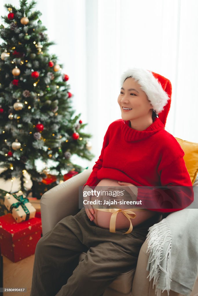 Donna incinta asiatica con cappello da Babbo Natale sorridente felice che mostra la pancia con fiocco seduta in soggiorno decorato con un albero di Natale.