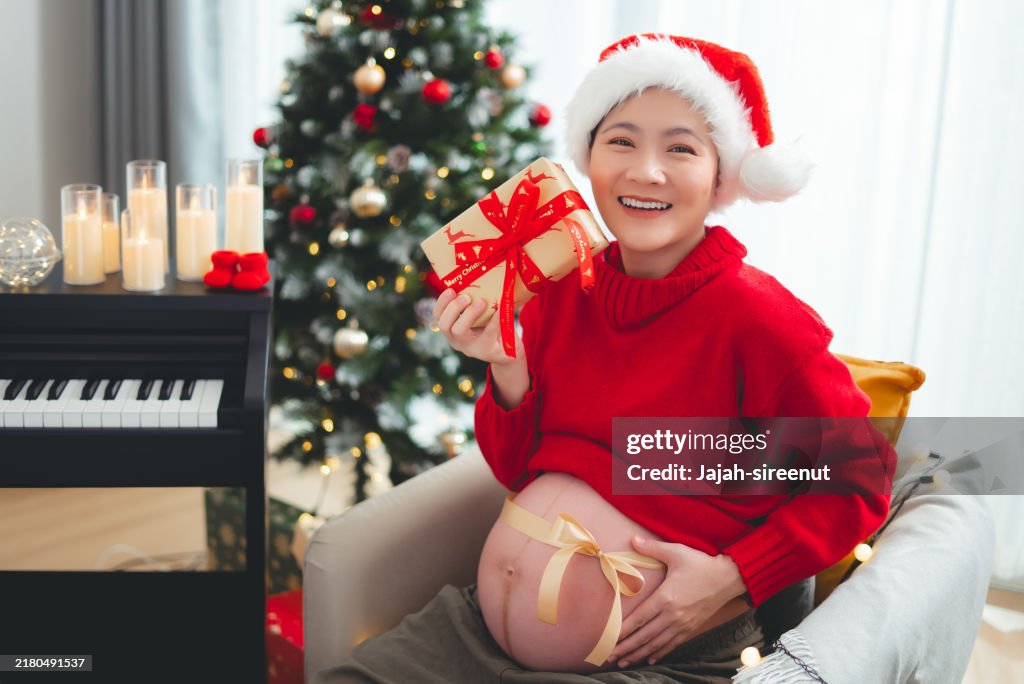 Donna incinta asiatica che indossa il cappello di Babbo Natale felice sorridente che mostra la pancia con fiocco e tiene in mano la scatola regalo seduta nel soggiorno decorato con un albero di Natale, guardando la macchina fotografica.