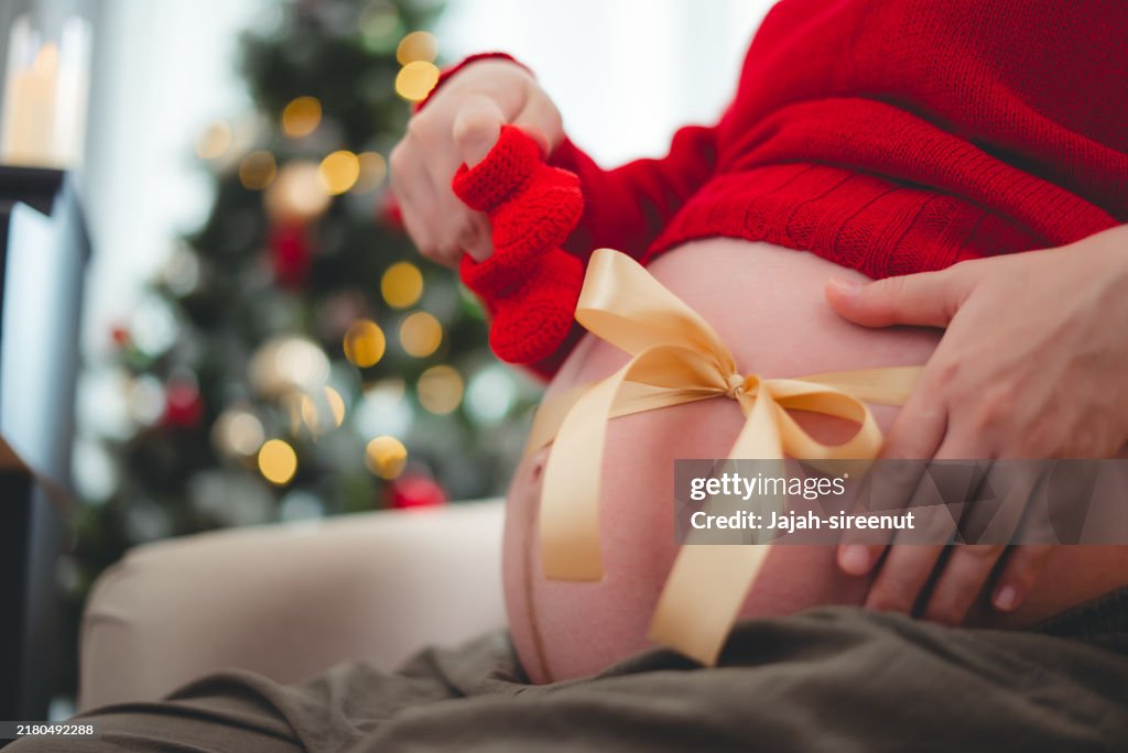 Close-up shot of pregnant woman showing belly with bow and holding crochet baby booties sitting in living room decorated with a Christmas tree.