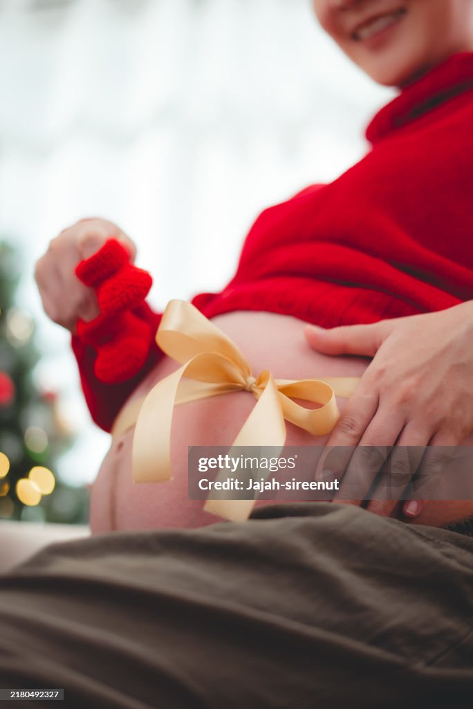 Close-up shot of pregnant woman showing belly with bow and holding crochet baby booties sitting in living room decorated with a Christmas tree.