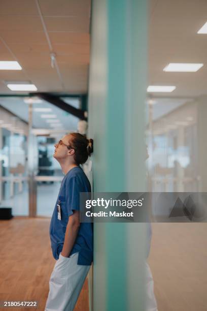 side view of tired non-binary healthcare expert leaning on wall in corridor at hospital - tired healthcare worker stock pictures, royalty-free photos & images