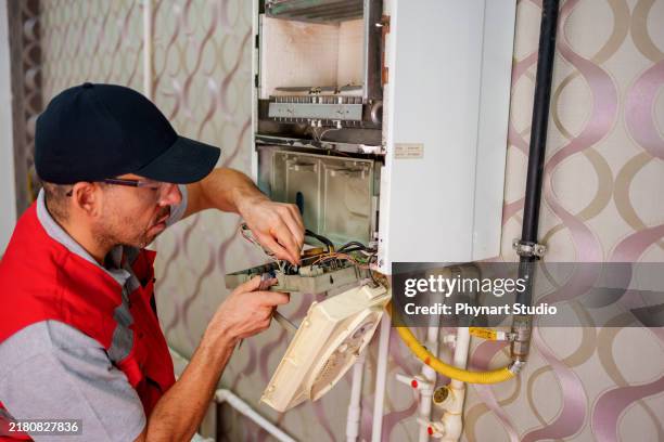 a technician in a red vest and hat carefully works on the internal components of a wall-mounted boiler. . - furnace stock pictures, royalty-free photos & images