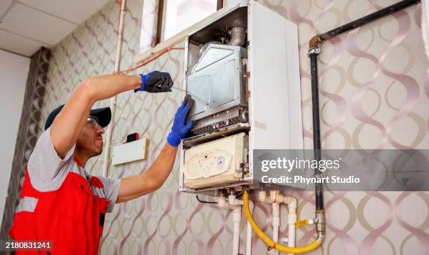 a technician in a red vest and hat carefully works on the internal components of a wall-mounted boiler. . - electric fire stock pictures, royalty-free photos & images