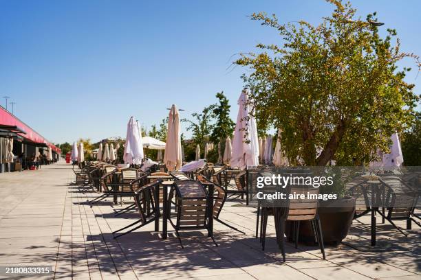 side view of a group of tables with wooden chairs and umbrellas in a courtyard with trees and no people around. - terraza fotografías e imágenes de stock