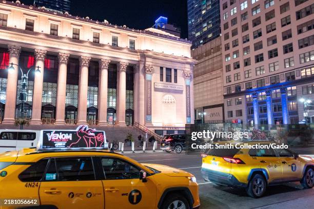 Illuminated facade of Moynihan Train Hall at night with yellow NYC taxis passing by, New York City, New York, October 20, 2024.