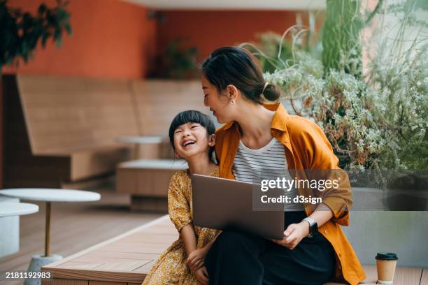 loving young asian mother and her daughter sitting in an outdoor cafe, using laptop together. they are enjoying time and smiling joyfully. mother-daughter bonding. digital native. family lifestyle with technology - digital native stock-fotos und bilder