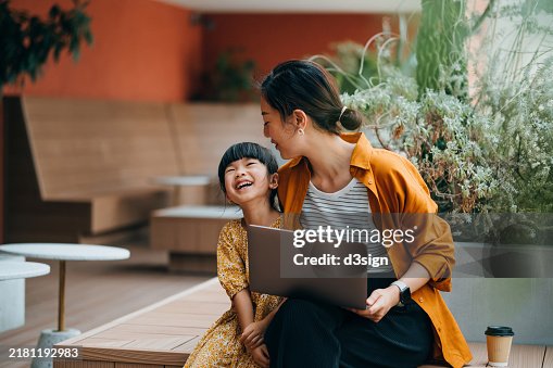 Loving young Asian mother and her daughter sitting in an outdoor cafe, using laptop together. They are enjoying time and smiling joyfully. Mother-daughter bonding. Digital native. Family lifestyle with technology Loving young Asian mother and her daughter sitting in an outdoor cafe, using laptop together. They are enjoying time and smiling joyfully. Mother-daughter bonding. Digital native. Family lifestyle with technology