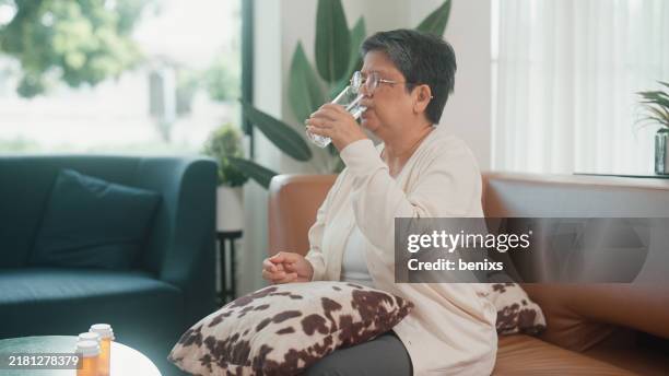 senior woman taking a pill with a glass of water in a bright and cozy living room - plastic water bottle open stock pictures, royalty-free photos & images