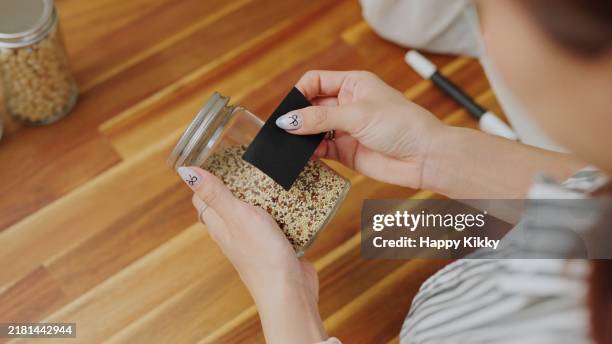 close-up hands labeling with black on a glass jar filled on a wooden counter for organized kitchen pantry storage ,zero waste lifestyle with green shopping,plastic free and sustainability at home - étiqueter photos et images de collection