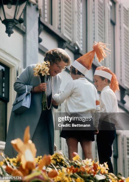 On the day of her 60th birthday, Queen Juliana of the Netherlands is presented with a bouquet of daffodils by two children, both wearing orange paper...