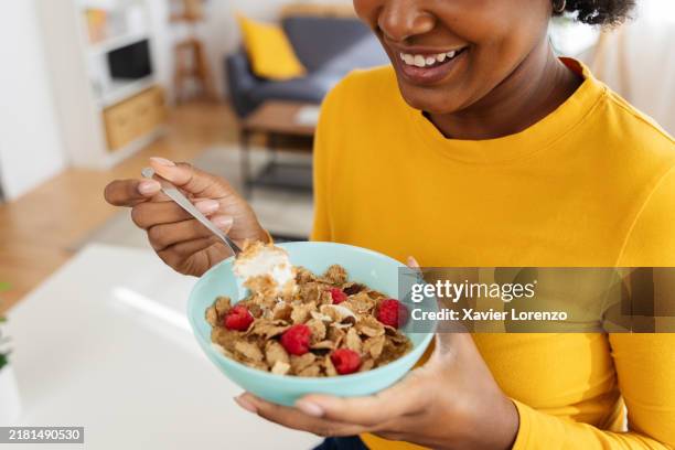 happy black woman eating breakfast cereal with raspberries and yogurt at home - fibra fotografías e imágenes de stock