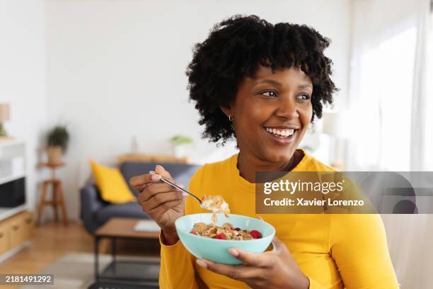 happy young black woman eating cereal with berries at home in living room - fibra fotografías e imágenes de stock