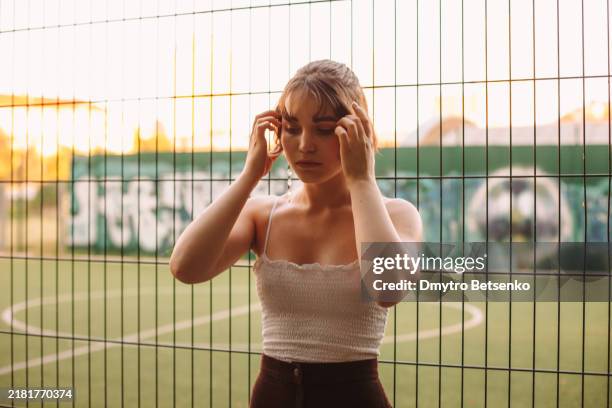 portrait of young woman standing by the fence against soccer field at campus - fobia imagens e fotografias de stock