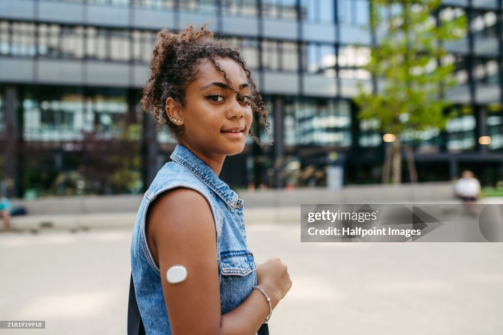 Arm with CGM sensor on it. Young diabetic woman in the city, walking on street, looking at camera.