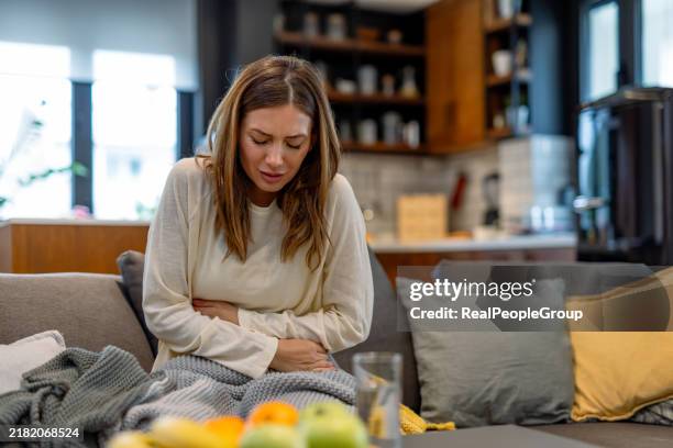 frau mit schmerzen, die den bauch auf dem wohnzimmersofa hält. - diarrhö stock-fotos und bilder