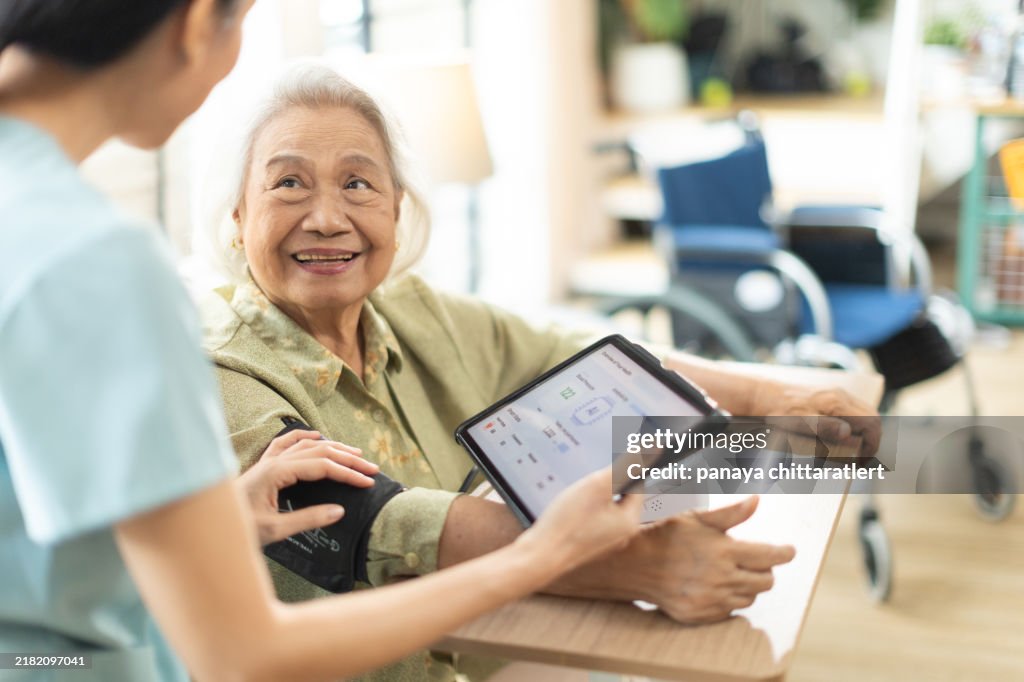 Woman asian nurse taking blood pressure of a senior woman patient at home.Home care healthcare professional using digital tablet