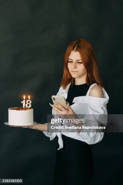 teenage girl with a smartphone in her hands holds a cake with burning candles in the shape of the number 18 on a black background - 18º aniversário imagens e fotografias de stock