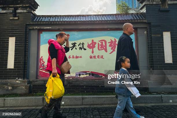 Grandparents escort their grandchild to a weekend educational class, passing by a wall displaying a slogan that reads “Building the Chinese Dream” on...