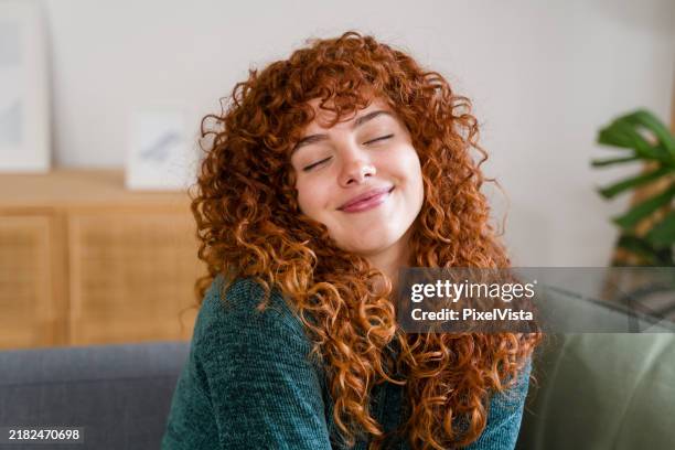 young woman smiling with eyes closed feeling relaxed on sofa - roodhoofd stockfoto's en -beelden