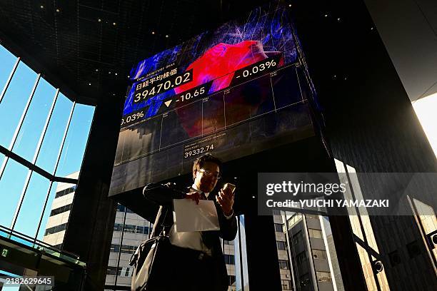 Man checks his phone as he walks past an electronic board displaying the numbers on the Tokyo Stock Exchange in Tokyo on November 7, 2024.