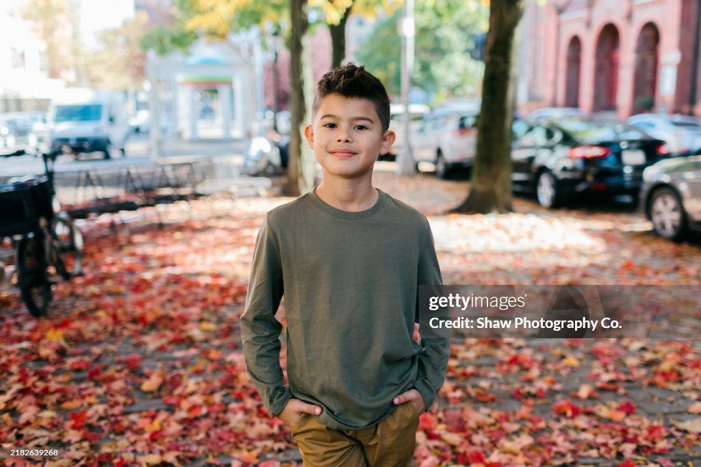 A mixed-race boy about 7-8 years old standing with his hands in his pocket on a leaf-lined street in New York City / Upper Manhattan / Inwood