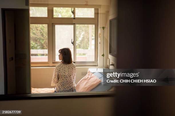 the back view of an asian female patient, seen through the reflection of glass, as she sits on the hospital bed contemplating her condition and future. - post operative stock pictures, royalty-free photos & images