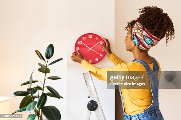 rear view of a brunette woman in casual clothes standing placing a clock on the wall of her apartment. - relógio de parede imagens e fotografias de stock