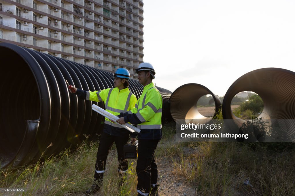 Two male structural experts inspect large drainage pipes while carrying a toolbox and a blueprint before sending it to pre-construction for the next process.