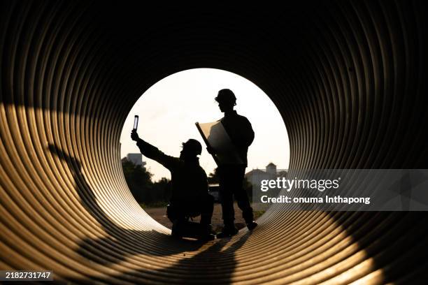 los trabajadores de la construcción y los ingenieros trabajan de manera eficiente dentro de una gran tubería de metal en un sitio de construcción. están construyendo un nuevo sistema de drenaje de agua por la noche, justo antes de la puesta del sol. - industria metálica fotografías e imágenes de stock