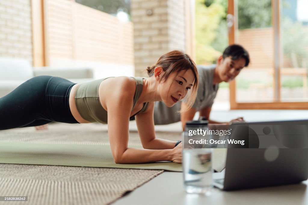 Young Asian couple taking online fitness class through laptop, working out together at home