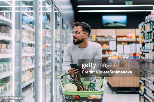 Cheerful man choosing products and using phone during grocery shopping in supermarket. Cheerful man choosing products and using phone during grocery shopping in supermarket.