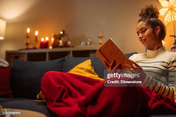 young woman reading a book on sofa at home during christmas time - vela equipamento de iluminação imagens e fotografias de stock