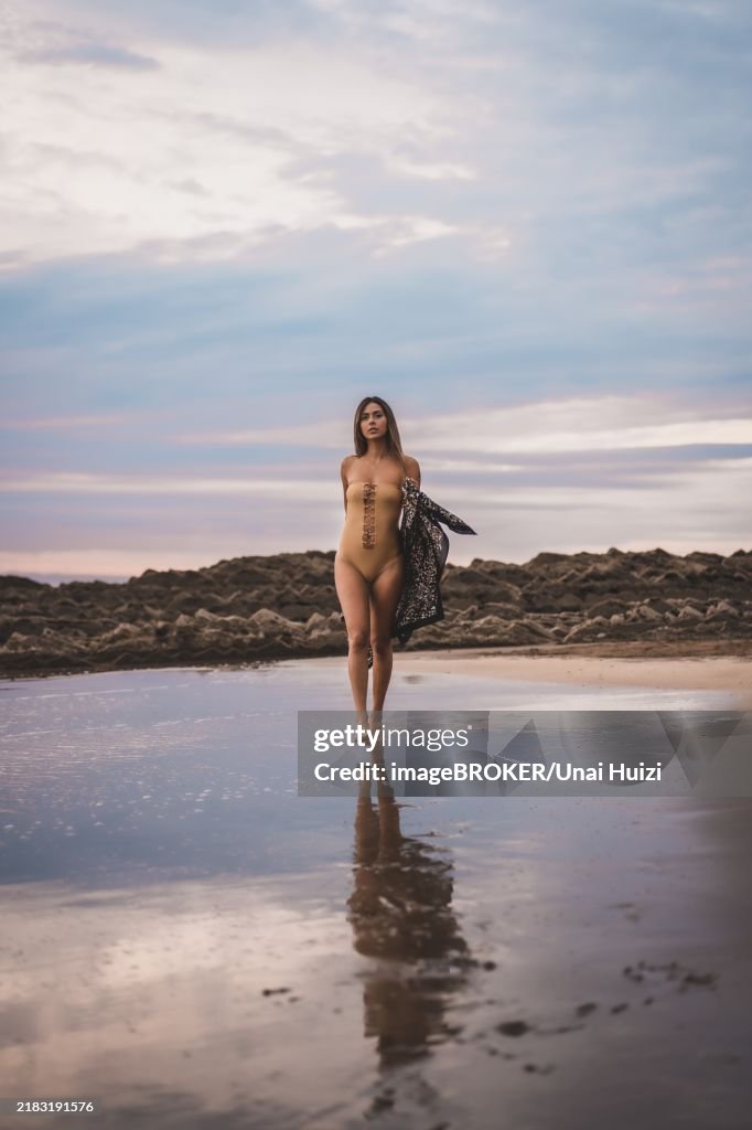 A brunette woman with a swimsuit and a pareo walking along the beach in summer in the cloudy sunset
