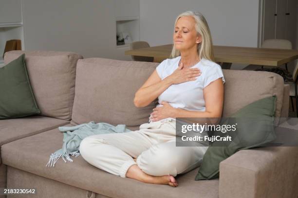 elderly woman meditating, doing breathing exercises sitting on sofa - hand on heart stock pictures, royalty-free photos & images
