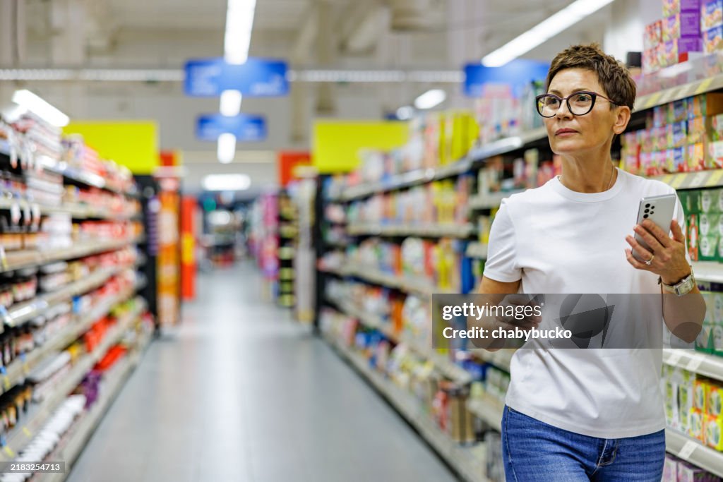 Beautiful lady searching grocery in supermarket following shopping list on smartphone