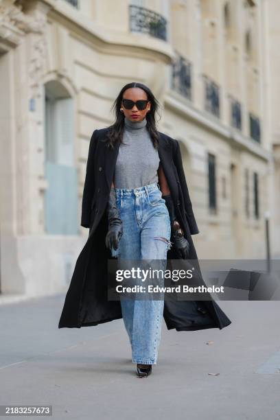 Emilie Joseph wears black sunglasses, silver earrings, a grey sleeveless turtleneck pullover, a black long coat, black leather gloves, dark brown...