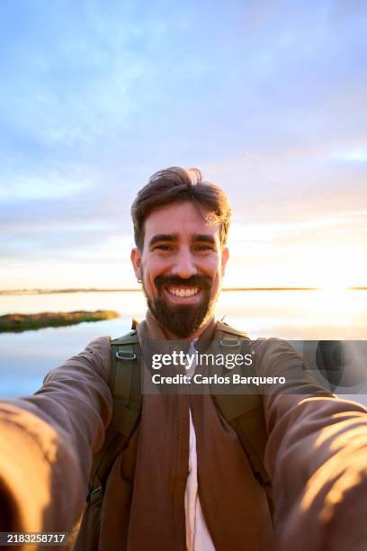 smiling man taking a selfie at sunset by a calm lake with scenic natural surrounding. - 35 mm stock pictures, royalty-free photos & images