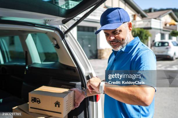 delivery man checking time while delivering parcels from car trunk - timely delivery stock pictures, royalty-free photos & images