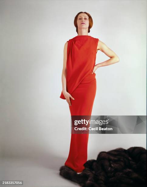 Russian ballerina, Maya Plisetskaya, wearing a red crepe chiffon gown by Gres. She stands on a brown fur that is draped along the floor.