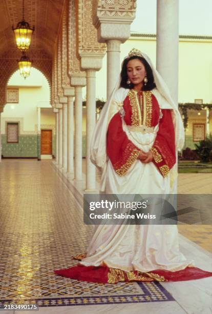 Princess Lalla Nouzha of Morocco wearing a gold encrusted red and white royal caftan in the palace courtyard on her wedding day.