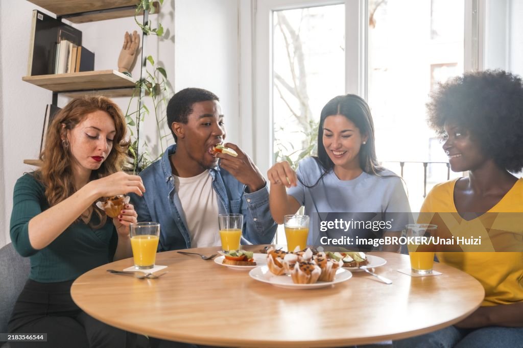Multiethnic friends at a breakfast with orange juice and muffins at home, detail cutting the toasts