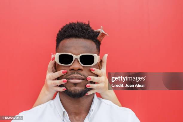 man with sunglasses poses dramatically in front of a red wall while a woman's hands touch his face - heteroseksueel koppel stockfoto's en -beelden