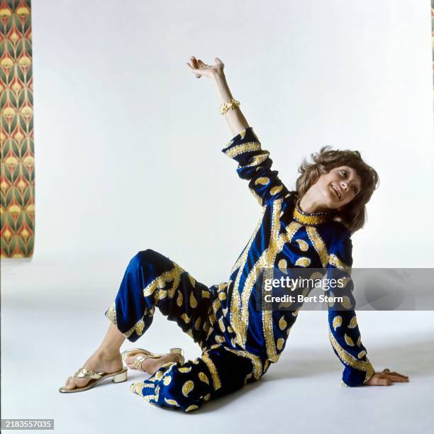 Model Loulou de la Falaise sits on a white studio backdrop with one knee up, leaning back on her left arm and extending her right arm into the air....