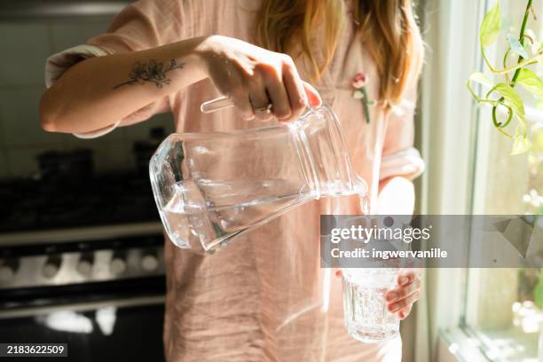 woman pouring water in a glass from a jug in the kitchen - sediento fotografías e imágenes de stock