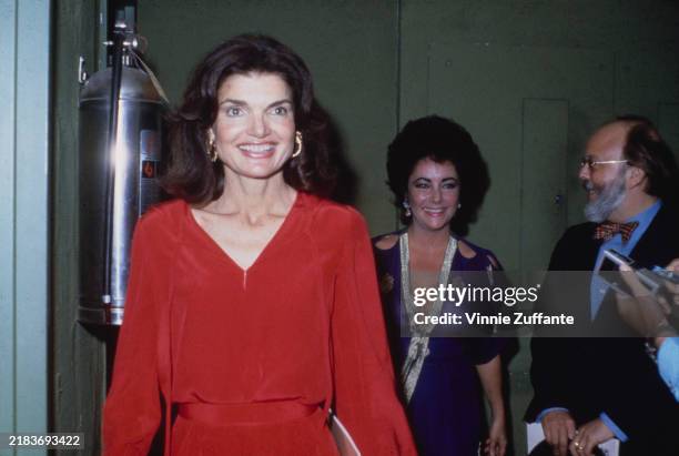 American socialite Jacqueline Kennedy Onassis, the former US First Lady, wearing a red v-neck outfit with gold hoop earrings, and British-American...